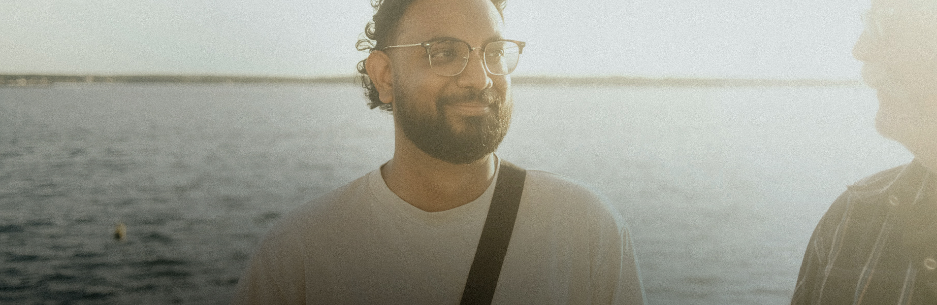 a person standing and talking to someone else. they're wearing glasses and a body of water is behind them. 